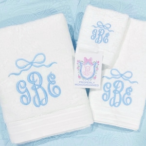 May include: White monogrammed towel set with light blue embroidered bow and initials. The set includes a large bath towel, a hand towel, and a washcloth. A tag reads "Properly Monogrammed".