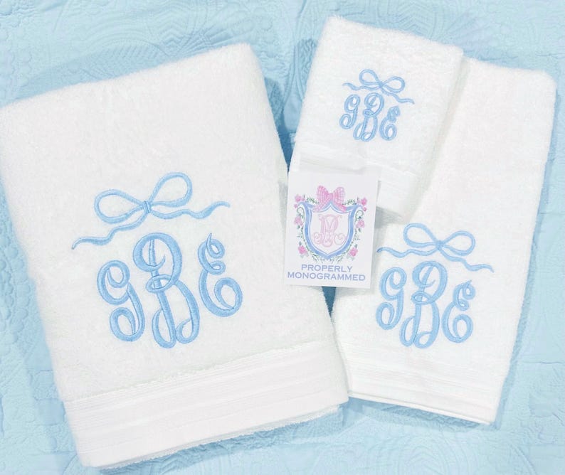 May include: White monogrammed towel set with light blue embroidered bow and initials. The set includes a large bath towel, a hand towel, and a washcloth. A tag reads "Properly Monogrammed".