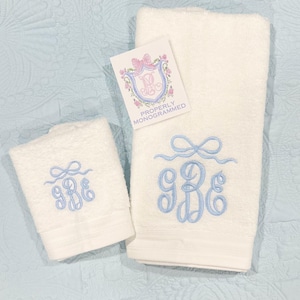 May include: Two white monogrammed towels with light blue embroidery. The towels have a bow design above the monogram. A small tag with a pink bow and floral design is visible. The text on the tag reads "Properly Monogrammed."