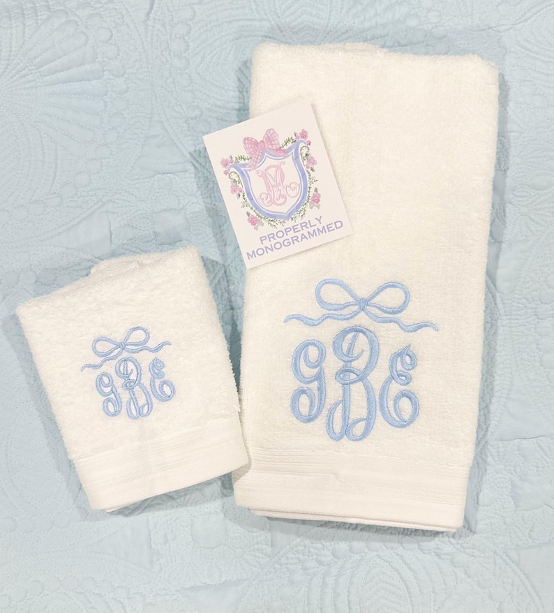 May include: Two white monogrammed towels with light blue embroidery. The towels have a bow design above the monogram. A small tag with a pink bow and floral design is visible. The text on the tag reads "Properly Monogrammed."