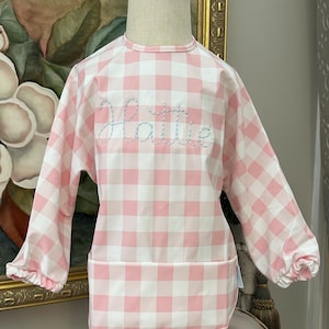 May include: A pink and white gingham bib with long sleeves. The bib features the name "Hattie" embroidered in light blue cursive script across the chest. The bib has a pocket at the bottom and elastic cuffs.