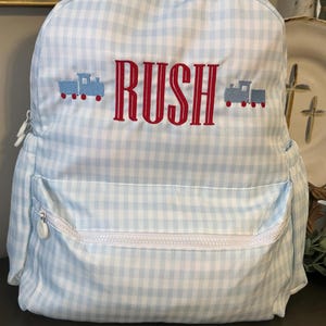 May include: A light blue and white gingham backpack with the name "RUSH" embroidered in red. The backpack features two blue train designs and a white zipper pocket. This backpack is perfect for school or travel.
