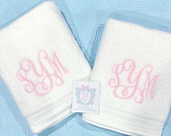 May include: Two white bath towels with pink embroidered initials. Each towel has a light gray stripe detail at the bottom. A small card with a floral crest and the words "Properly Monogrammed" is between the towels.