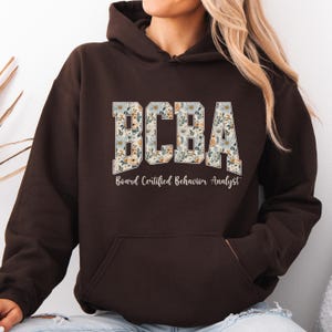 May include: A dark brown hoodie with the letters "BCBA" in a floral pattern across the chest. Below the letters, the text reads "Board Certified Behavior Analyst" in a simple font. The hoodie has a front pocket and a hood.