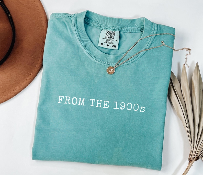 Op de afbeelding: Een turquoise Comfort Colors t-shirt met de tekst "FROM THE 1900s" in het wit. Een gouden ketting met een hanger ligt op het shirt. Een bruine hoed en een gedroogd palmblad zijn op de achtergrond.