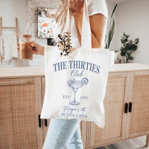 Peut inclure: Sac fourre-tout cr&egrave;me avec l'inscription bleue "THE THIRTIES Club". En dessous, une illustration bleue d'un cocktail. Le sac porte &eacute;galement les mentions "EST 1995" et "Morgan's 30th PUNTA CANA".