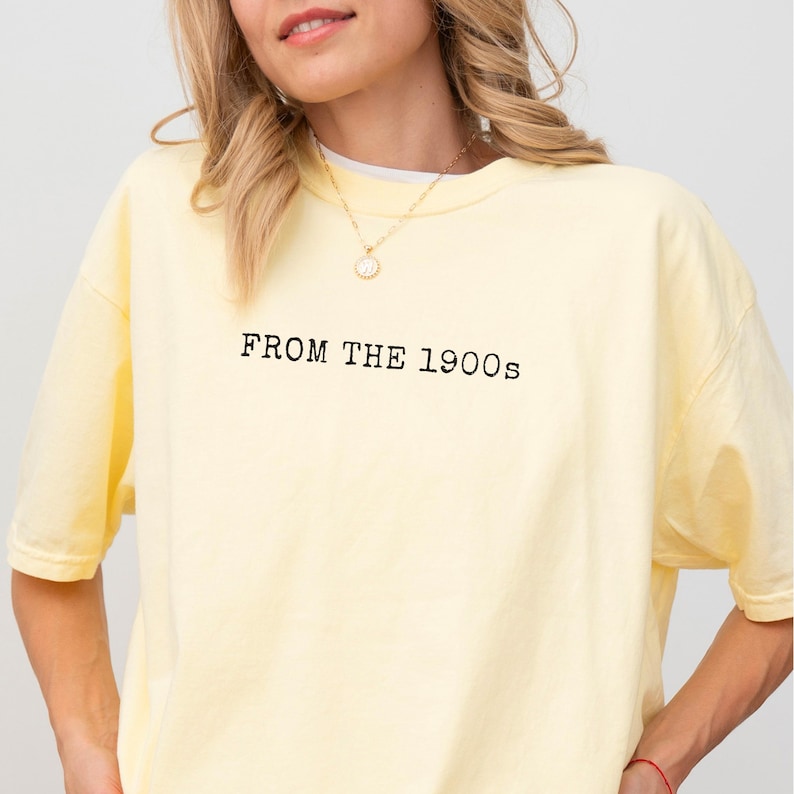 Op de afbeelding: Lichtgele t-shirt met de tekst "FROM THE 1900s" in het zwart gedrukt. Het shirt heeft een ronde hals en een relaxte pasvorm. Een gouden ketting is zichtbaar.