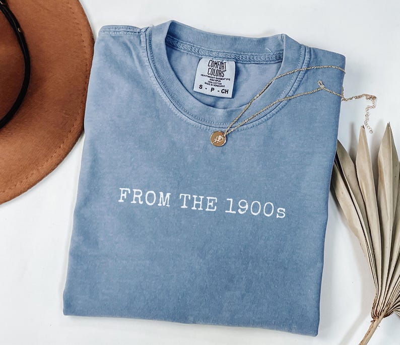 Op de afbeelding: Een blauw T-shirt met de tekst "FROM THE 1900s" in het wit. Een gouden ketting met een hanger ligt over het shirt. Een bruine hoed is zichtbaar in de linkerbovenhoek.