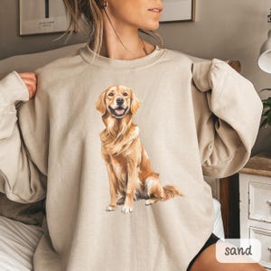 以下が含まれることがあります： ゴールデンレトリバー犬の詳細なイラストが描かれた、砂色のスウェットシャツ。犬は幸せそうな表情で座った姿勢で描かれています。「sand」の文字が右下にプリントされています。スウェットシャツは柔らかい素材で作られています。