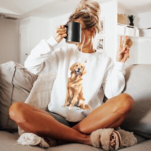 以下が含まれることがあります： ゴールデンレトリバー犬の水彩イラストが描かれた白いスウェットシャツ。犬は幸せそうな表情で座っています。スウェットシャツはカジュアルなスタイルで、普段使いに適しています。