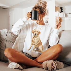 以下が含まれることがあります： 前面にゴールデンレトリバーの犬が座っている水彩画のイラストが描かれた白いスウェットシャツ。ソファに座っている人が着ています。