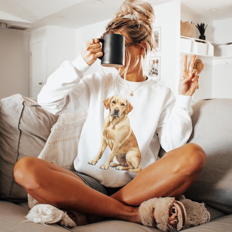以下が含まれることがあります： 前面にゴールデンレトリバーの犬が座っている水彩画のイラストが描かれた白いスウェットシャツ。ソファに座っている人が着ています。