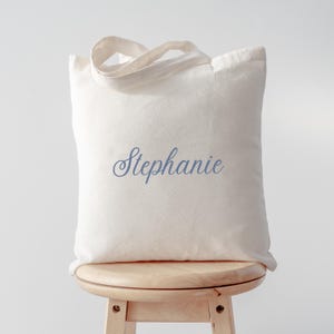 Peut inclure: Un sac fourre-tout cr&egrave;me avec le nom "Stephanie" en cursive bleue. Le sac a un design simple avec deux poign&eacute;es et est pos&eacute; sur un tabouret en bois.