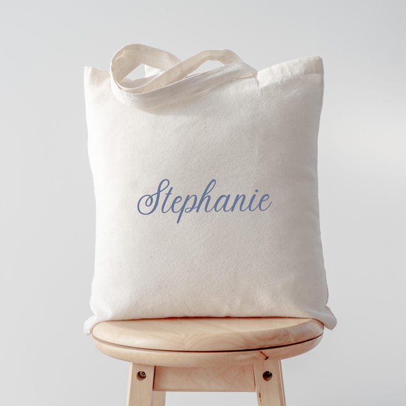 Peut inclure: Un sac fourre-tout cr&egrave;me avec le nom "Stephanie" en cursive bleue. Le sac a un design simple avec deux poign&eacute;es et est pos&eacute; sur un tabouret en bois.