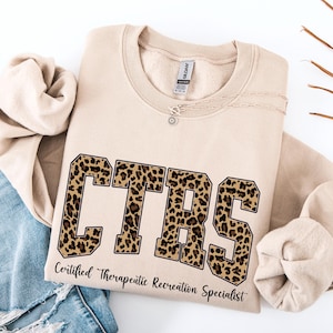 May include: Beige sweatshirt with the letters "CTRS" in a leopard print design. The text "Certified Therapeutic Recreation Specialist" is printed below the letters. A silver necklace is draped across the sweatshirt.