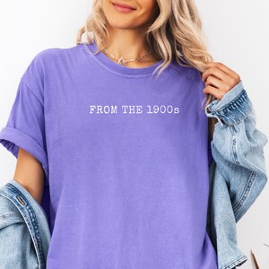Op de afbeelding: Een lavendelkleurig T-shirt met de tekst "FROM THE 1900s" in het wit gedrukt. Het shirt heeft opgerolde mouwen en wordt gecombineerd met een spijkerjasje. De algehele stijl is casual en relaxed.