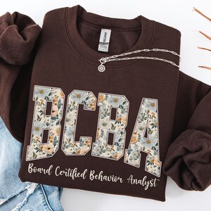 May include: A dark brown sweatshirt with the letters "BCBA" in a floral design. Below the letters, the text reads "Board Certified Behavior Analyst". A silver necklace with a pendant is draped across the sweatshirt.