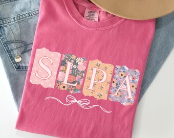 Puede incluir: Camiseta rosa con la palabra "SLPA" escrita en letras con estampado floral. Cada letra está rodeada de un borde blanco y tiene un efecto de papel rasgado. Una cinta blanca con un lazo se encuentra debajo de las letras.