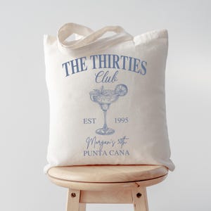 Peut inclure: Sac fourre-tout en toile &eacute;crue avec texte bleu clair et un dessin de cocktail. Le texte indique "THE THIRTIES Club" au-dessus d'une illustration de margarita, avec "EST 1995" et "Morgan's 30th PUNTA CANA" en dessous.