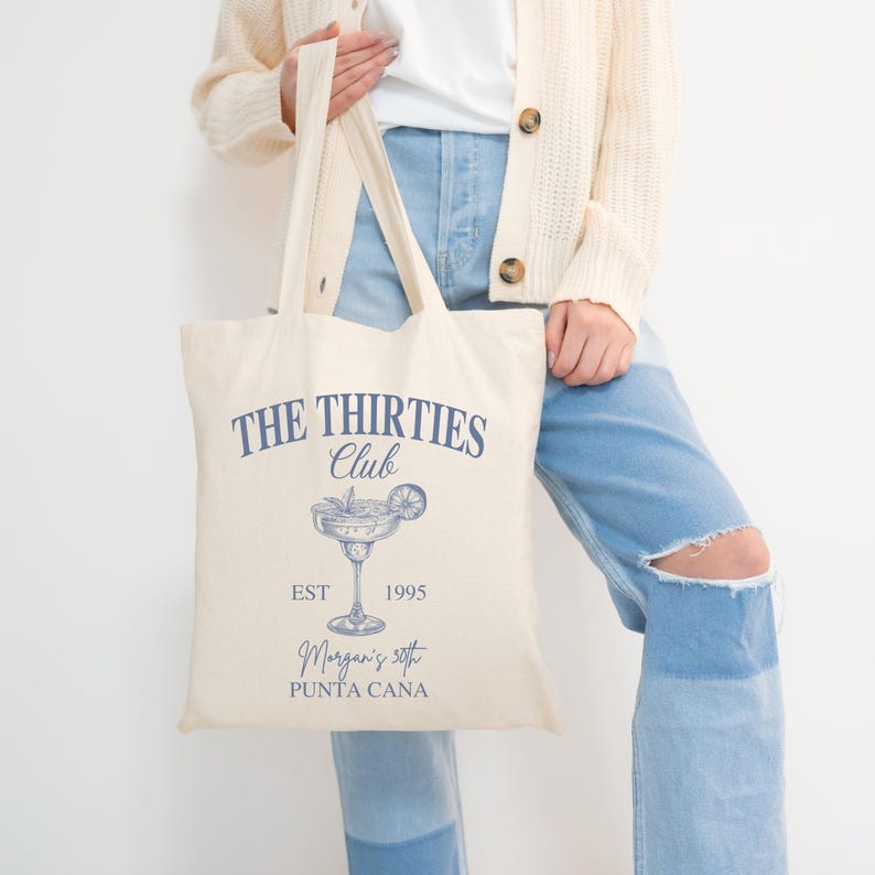 Peut inclure: Sac fourre-tout cr&egrave;me avec le texte "THE THIRTIES Club" en bleu marine. En dessous, une illustration de cocktail et les mots "EST 1995". Le bas du sac indique "Morgan's 50th PUNTA CANA". Le sac a deux anses cr&egrave;me.