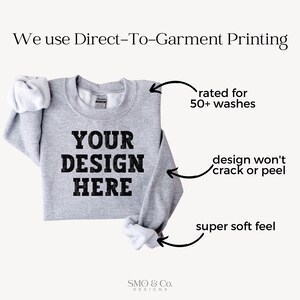 以下が含まれることがあります： 黒い文字で「YOUR DESIGN HERE」と書かれたグレーのスウェットシャツ。画像は「50回以上の洗濯に耐える」、「デザインがひび割れたり剥がれたりしない」、「非常にソフトな肌触り」などの特徴を強調しています。「We use Direct-To-Garment Printing」というテキストも表示されています。