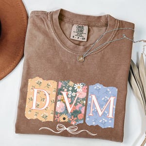 May include: A brown t-shirt with a floral design that spells out "DVM". The shirt is made of a soft, comfortable material and has a distressed look.