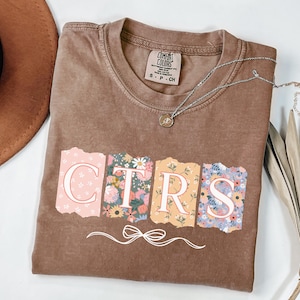 May include: A brown t-shirt with the word "CTRS" in a floral design. The letters are in a torn paper style with a bow graphic below. A silver necklace with a small pendant is on the shirt.