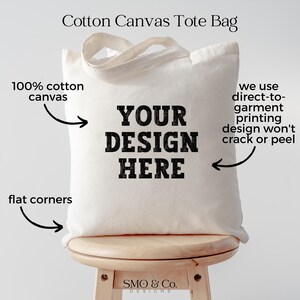 Peut inclure: Un sac fourre-tout en toile de coton cr&egrave;me avec le texte "YOUR DESIGN HERE" en lettres noires grasses. Le sac a des coins plats et une poign&eacute;e. Le texte sur le sac indique qu'il est fait de toile 100% coton et utilise l'impression directe sur v&ecirc;tement.