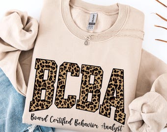 May include: Beige sweatshirt with the letters "BCBA" in a leopard print design. Below the letters is the text "Board Certified Behavior Analyst" in a script font. A delicate necklace with a small pendant is visible.