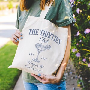 Peut inclure: Sac fourre-tout cr&egrave;me avec le texte "THE THIRTIES Club" et une illustration de margarita. Le sac comprend &eacute;galement le texte "EST 1995" et "Morgan's 30th PUNTA CANA". Le sac a un design simple avec une bandouli&egrave;re.