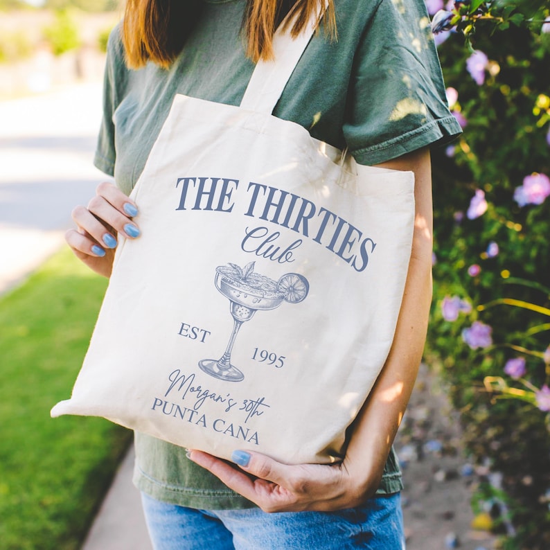 Peut inclure: Sac fourre-tout cr&egrave;me avec le texte "THE THIRTIES Club" et une illustration de margarita. Le sac comprend &eacute;galement le texte "EST 1995" et "Morgan's 30th PUNTA CANA". Le sac a un design simple avec une bandouli&egrave;re.