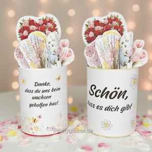 Op de afbeelding: Twee witte cilindrische containers met bloemen- en vlindermotieven, elk met decoratieve papieren bloemen en een hartvormige opstelling van rode rozen. Op een container staat "Danke, dass du uns beim wachsen geholfen hast."