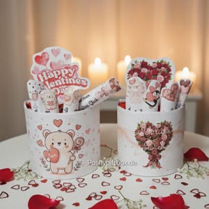以下が含まれることがあります： バレンタインデーをテーマにしたデザインの2つの円筒形のギフトボックス。1つはハートを持ったクマと「Happy Valentines Day」の文字が特徴です。もう1つはバラの花束です。赤いバラの花びらとハートの紙吹雪が散らばっています。
