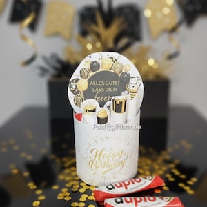 May include: A white gift box with gold "Happy Birthday" text and a decorative topper with balloons and the text "Alles Gute! Lass Dich Feier". Two red and white chocolate bars are in front of the box, with gold confetti scattered around.