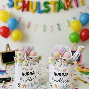 K&ouml;nnte beinhalten: Zwei wei&szlig;e zylindrische Beh&auml;lter mit dem Text "HURRA! Endlich Schulkind" sind mit Bleistiften, Radiergummis und Luftballondekorationen gef&uuml;llt. Im Hintergrund befindet sich ein Banner mit der Aufschrift "SCHULSTART!" und bunte Luftballons.