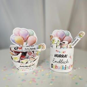 K&ouml;nnte beinhalten: Zwei wei&szlig;e Beh&auml;lter mit dem Text "HURRA! Endlich Schulkind" und bunten Konfetti. Einer ist eimerf&ouml;rmig, der andere zylindrisch. Beide haben Ballon- und Cartoon-Charakter-Dekorationen.