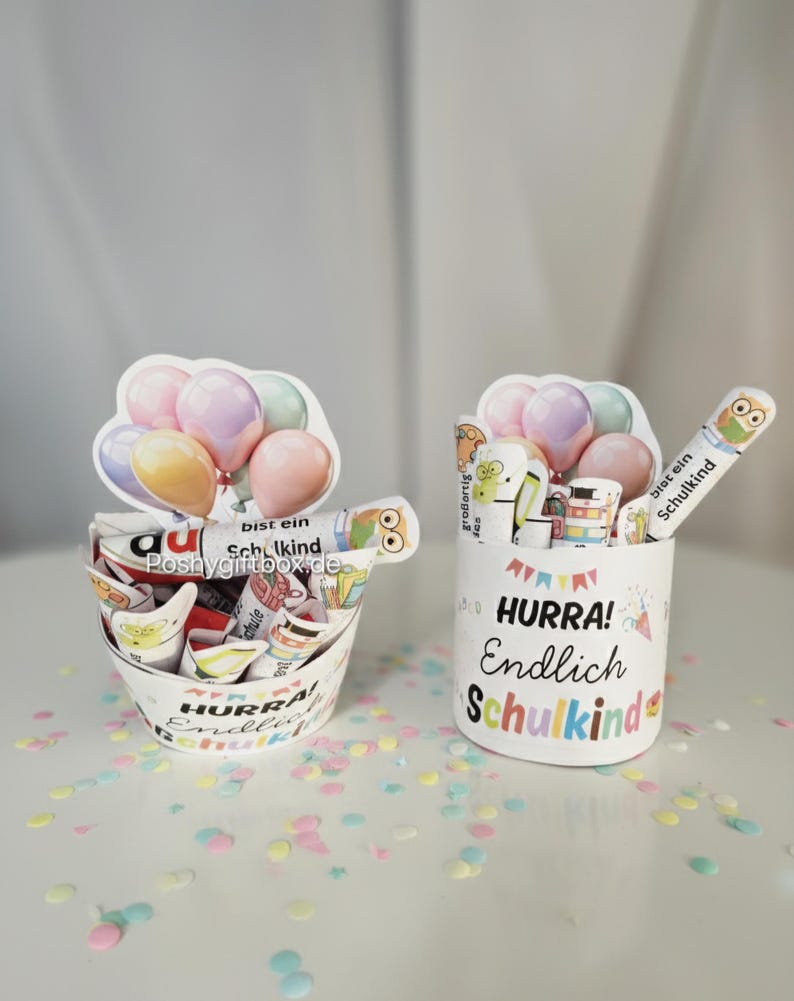 K&ouml;nnte beinhalten: Zwei wei&szlig;e Beh&auml;lter mit dem Text "HURRA! Endlich Schulkind" und bunten Konfetti. Einer ist eimerf&ouml;rmig, der andere zylindrisch. Beide haben Ballon- und Cartoon-Charakter-Dekorationen.