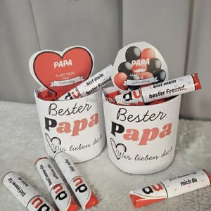Può includere: Due tazze bianche con le parole "Bester Papa" in nero e rosa, riempite di caramelle e decorazioni a forma di cuore. Le tazze hanno le parole "Wir lieben dich" sotto. Diverse caramelle con testo tedesco sono sparse.