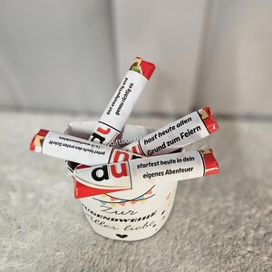 May include: A white ceramic cup filled with wrapped chocolate bars. The cup has text in German and a colorful design. The chocolate bars have red and white packaging with German text. The cup is on a soft, light-colored surface.
