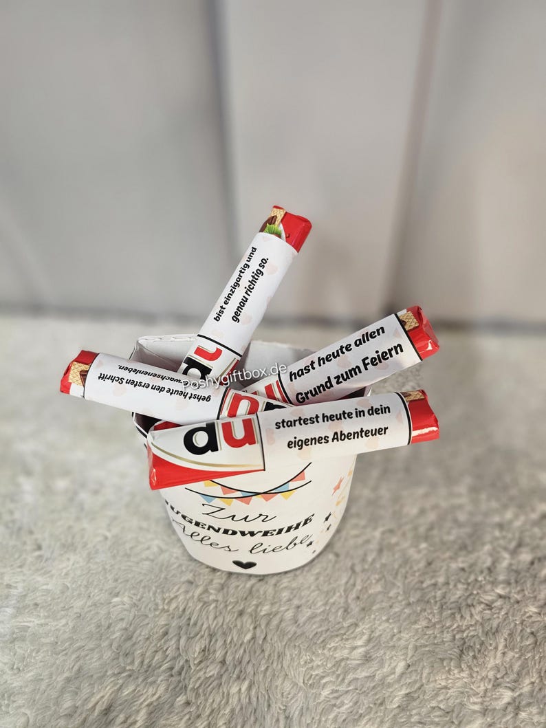 May include: A white ceramic cup filled with wrapped chocolate bars. The cup has text in German and a colorful design. The chocolate bars have red and white packaging with German text. The cup is on a soft, light-colored surface.
