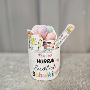 K&ouml;nnte beinhalten: Ein wei&szlig;er zylindrischer Beh&auml;lter mit den Worten "HURRA! Endlich Schulkind" in bunten Buchstaben. Der Beh&auml;lter ist mit dekorativen Gegenst&auml;nden gef&uuml;llt, darunter Luftballons, ein Bleistift mit Eulenmotiv und andere schulische Verzierungen.