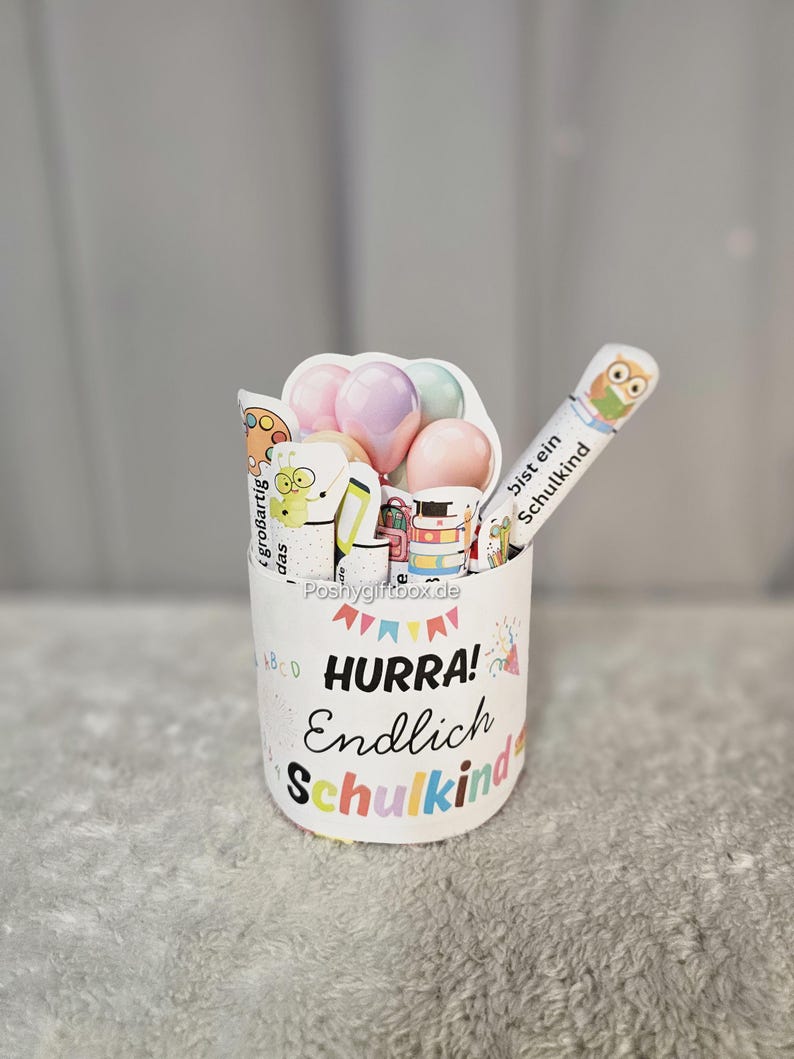 K&ouml;nnte beinhalten: Ein wei&szlig;er zylindrischer Beh&auml;lter mit den Worten "HURRA! Endlich Schulkind" in bunten Buchstaben. Der Beh&auml;lter ist mit dekorativen Gegenst&auml;nden gef&uuml;llt, darunter Luftballons, ein Bleistift mit Eulenmotiv und andere schulische Verzierungen.