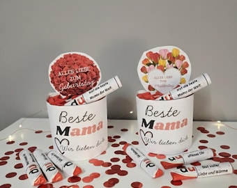May include: Two white cylindrical containers with the text "Beste Mama" and "Wir lieben dich" in pink and black. Each container has a floral topper and several wrapped candies. Red confetti is scattered around.