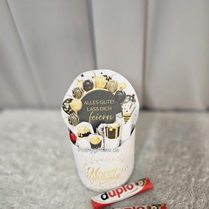May include: A white gift box with gold accents and the words "Happy Birthday" in gold script. The box contains a round card with the words "Alles Gute! Lass Dich feiern" and decorative balloons. Two "duplo" chocolate bars are in front of the box.