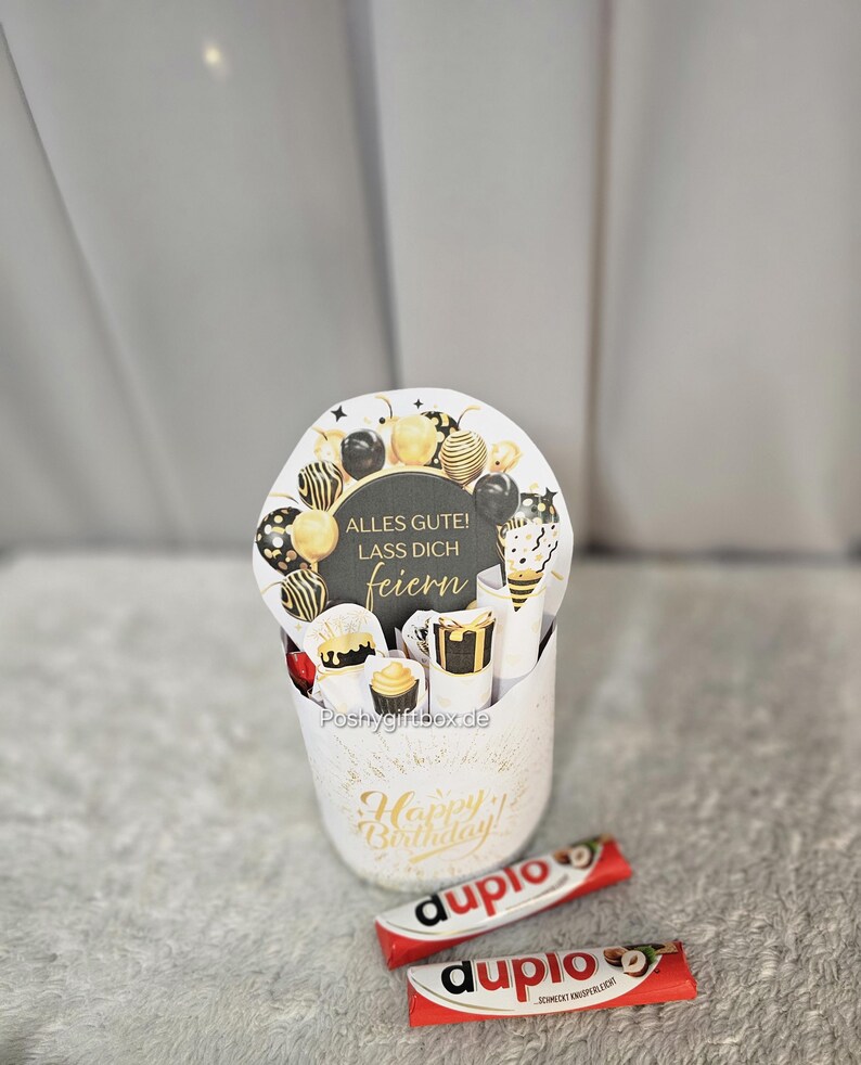 May include: A white gift box with gold accents and the words "Happy Birthday" in gold script. The box contains a round card with the words "Alles Gute! Lass Dich feiern" and decorative balloons. Two "duplo" chocolate bars are in front of the box.