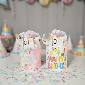 Könnte beinhalten: Zwei zylindrische Geburtstagsgeschenkboxen mit farbenfrohem "Happy Birthday"-Text und Illustrationen. Eine Box hat ein pastellfarbenes Regenbogen-Design, die andere einen weißen Hintergrund. Beide sind mit Leckereien gefüllt und mit dekorativen Elementen versehen.
