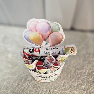 K&ouml;nnte beinhalten: Ein wei&szlig;er Papiergeschenkkorb mit farbenfrohem Text und Illustrationen, gef&uuml;llt mit verpackten S&uuml;&szlig;igkeiten und einem dekorativen ballonf&ouml;rmigen Aufsatz. Der Korb tr&auml;gt die Aufschrift "HURRA!" und "Schulkind".