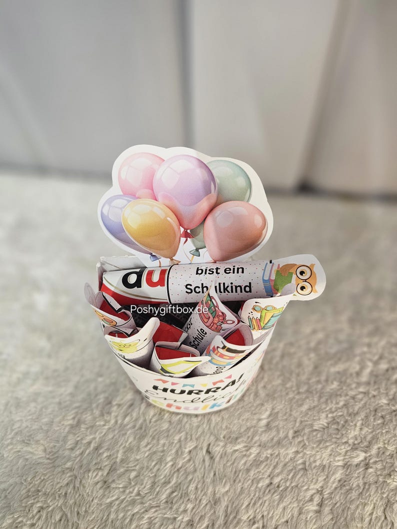 K&ouml;nnte beinhalten: Ein wei&szlig;er Papiergeschenkkorb mit farbenfrohem Text und Illustrationen, gef&uuml;llt mit verpackten S&uuml;&szlig;igkeiten und einem dekorativen ballonf&ouml;rmigen Aufsatz. Der Korb tr&auml;gt die Aufschrift "HURRA!" und "Schulkind".