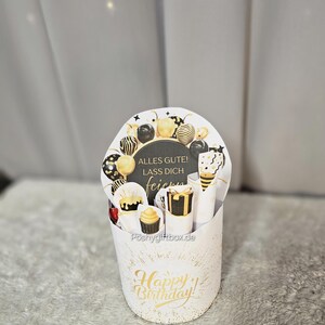 May include: A white gift box with gold accents and the words "Happy Birthday!" in gold script. The box contains a decorative card with balloons and the text "Alles Gute! Lass dich feiern". Inside are small gifts and treats.