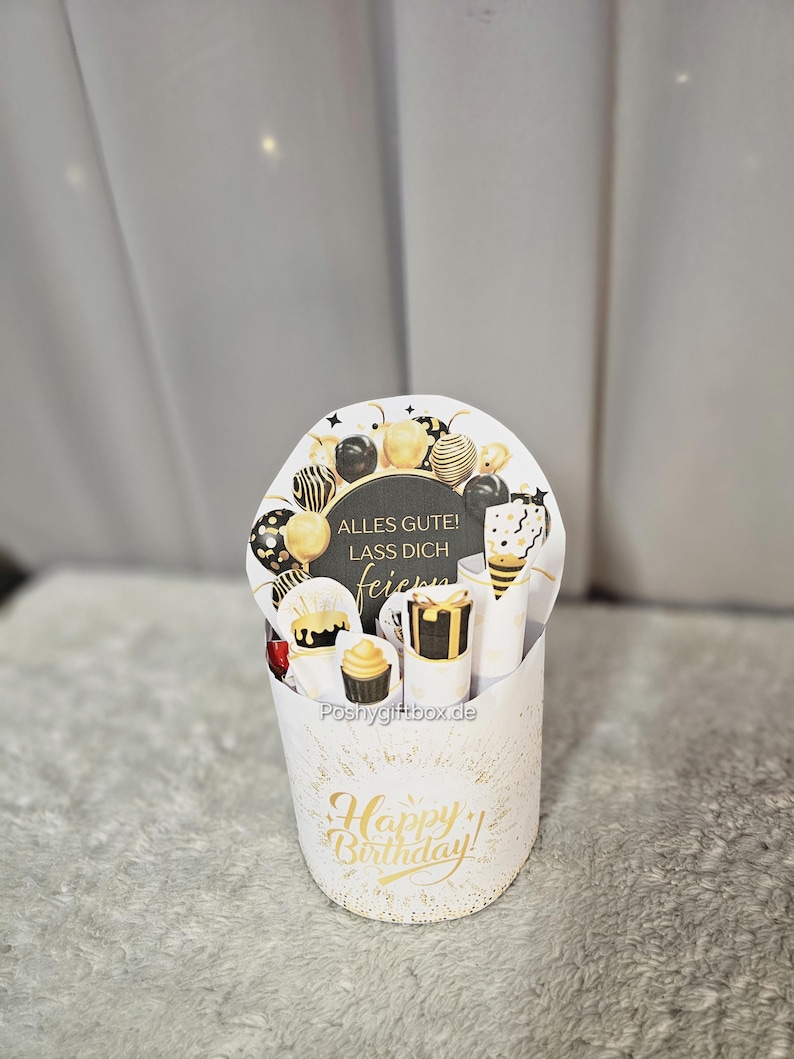 May include: A white gift box with gold accents and the words "Happy Birthday!" in gold script. The box contains a decorative card with balloons and the text "Alles Gute! Lass dich feiern". Inside are small gifts and treats.